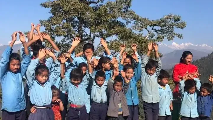 Primary school students raising their hands outdoors, with mountains visible behind them